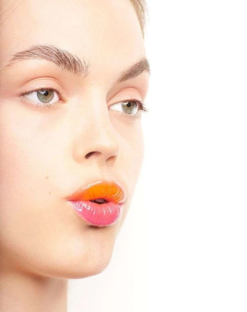 Close-up studio shot of a fair-skinned adult female with orange-pink ombre lips looking to the side against a plain white backdrop.