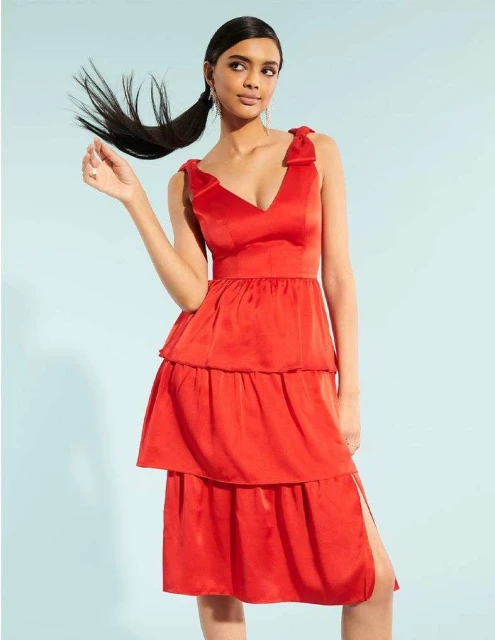 A South Asian woman poses in a red sleeveless tiered dress against a light blue backdrop, creating a vibrant fashion portrait.