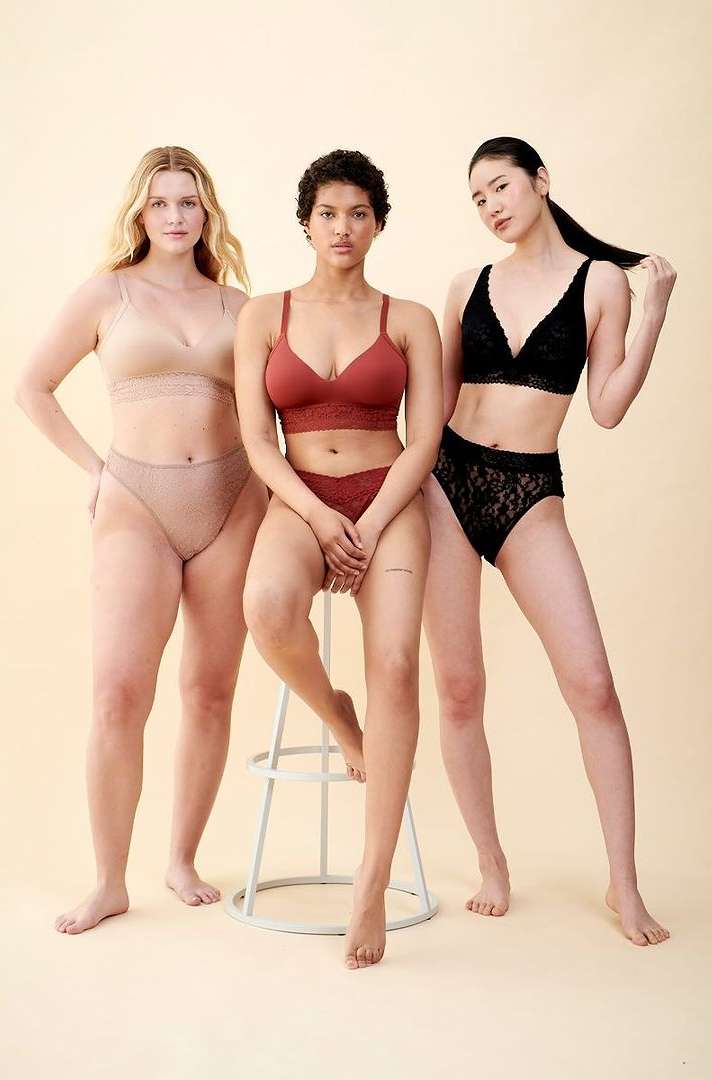 Three women pose in bras and underwear against a neutral background, showcasing fashion apparel in a studio setting with soft lighting.