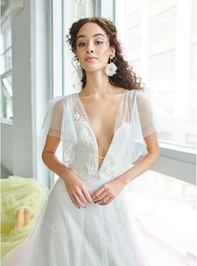 Elegant woman with curly hair poses in a white dress with sheer overlay and flower earrings, standing indoors by a window.