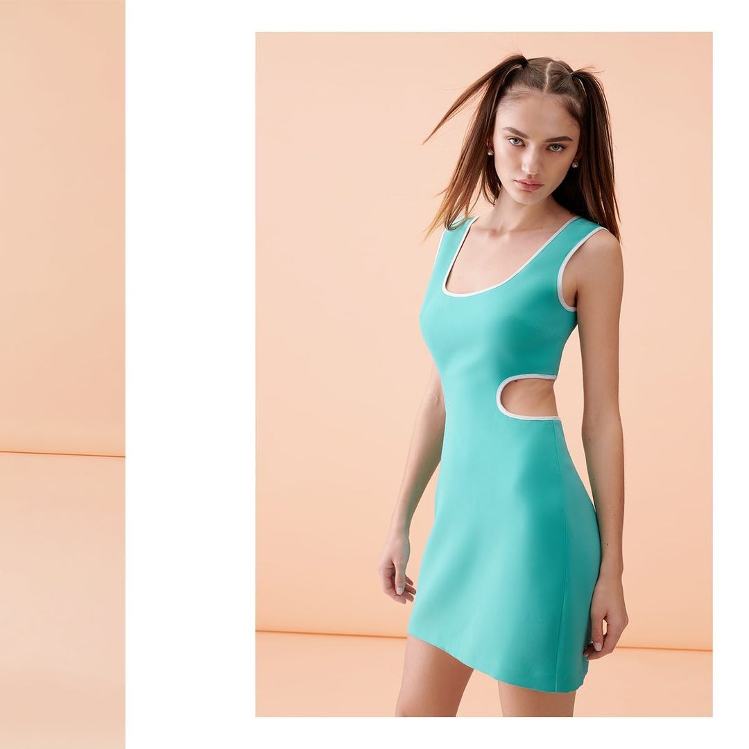 A woman poses in a studio wearing a sleeveless, short, mint green dress with white trim and two pigtails, set against a solid peach backdrop.