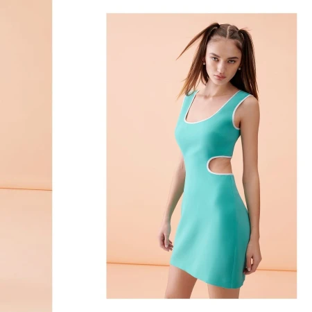 A woman poses in a studio wearing a sleeveless, short, mint green dress with white trim and two pigtails, set against a solid peach backdrop.