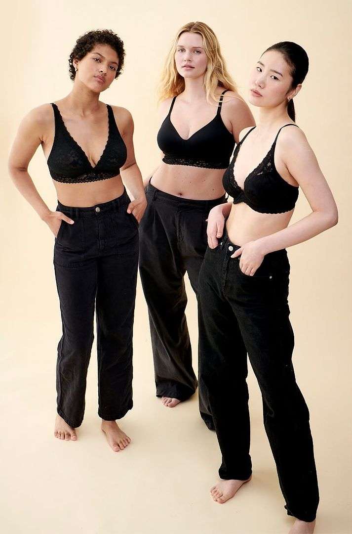 Three women with diverse ethnicities pose indoors wearing black lingerie tops and dark pants, showcasing casual fashion against a neutral backdrop.