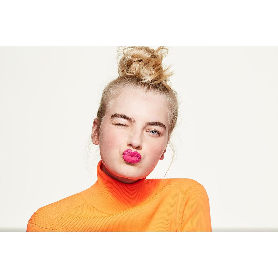 Close-up studio shot of a fair-skinned young adult female with an orange top, blonde hair, and bright pink lipstick winking with pursed lips.