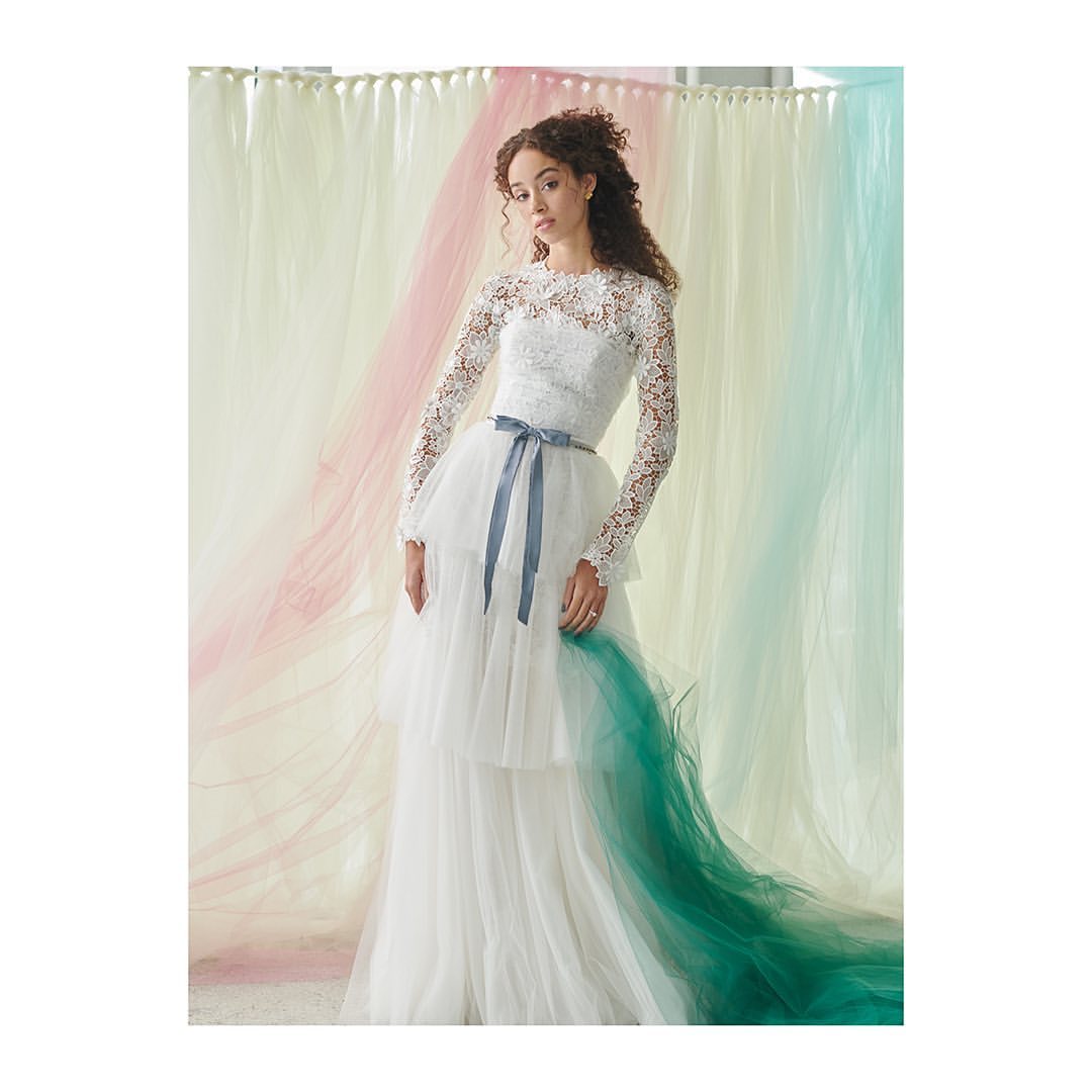 Woman wearing a lace long-sleeved wedding dress with a tiered skirt and blue sash stands in front of a pastel background.
