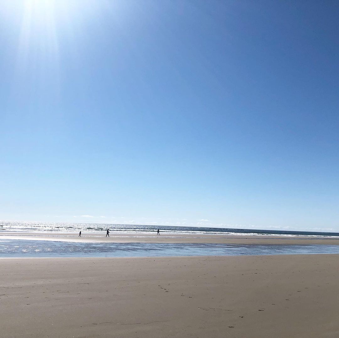 Wide angle shot of a expansive sandy beach and ocean under a bright blue sky with a few distant figures walking.