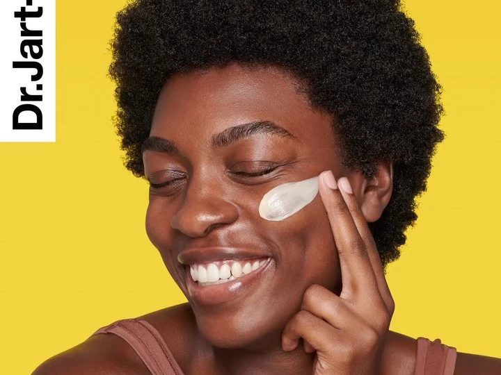 Close-up of a smiling Black woman applying cream to her face, set against a yellow background with brand logo.