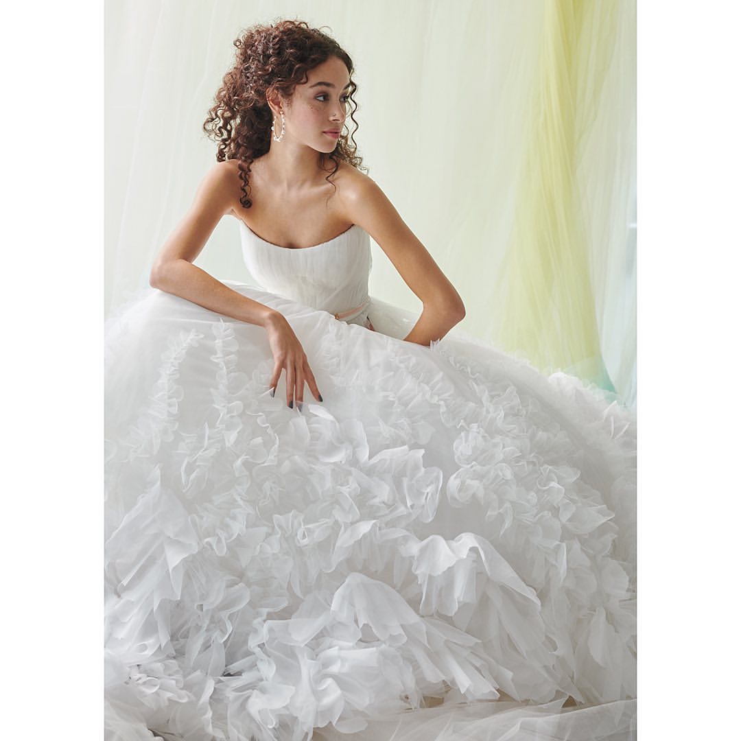 Brunette woman in a strapless wedding dress with a full, layered skirt, sitting in a light-filled studio with a translucent backdrop.