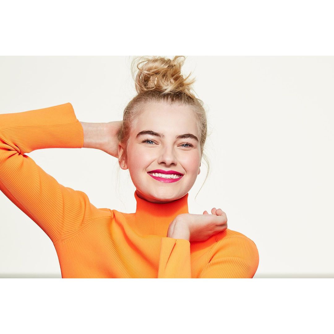 Blonde woman in an orange turtleneck sweater smiles in a studio portrait. Her hair is in a messy bun on top of her head.