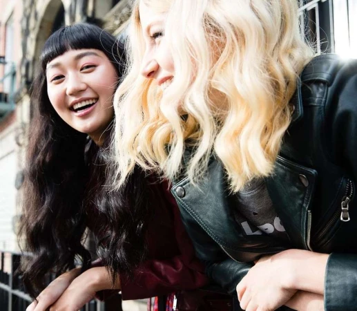 Two smiling young women leaning on a railing outdoors, one with dark hair and bangs and one with blonde wavy hair wearing a black leather jacket.