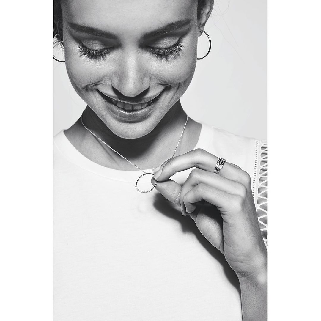 Close-up studio shot of a smiling woman in a white shirt holding a round silver pendant necklace and wearing hoop earrings and rings.