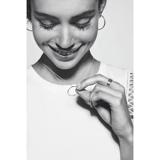 Close-up studio shot of a smiling woman in a white shirt holding a round silver pendant necklace and wearing hoop earrings and rings.