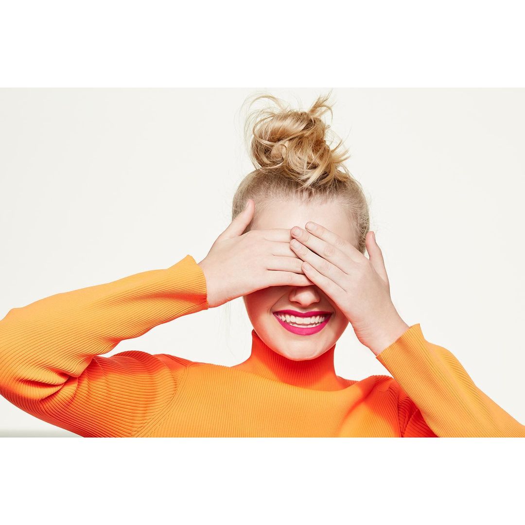 A cheerful young woman with blonde hair in a bun covers her eyes while wearing a bright orange turtleneck sweater against a white background.