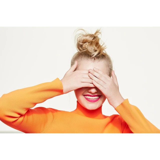 A cheerful young woman with blonde hair in a bun covers her eyes while wearing a bright orange turtleneck sweater against a white background.