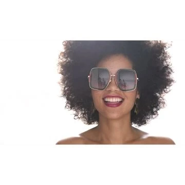 A smiling African American woman with curly hair wears oversized sunglasses and hoop earrings against a bright, white background in a headshot.