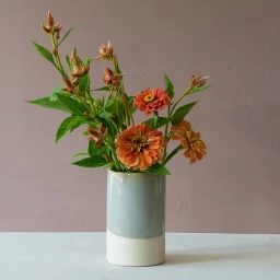 Orange flowers and green foliage arranged in a two-tone ceramic vase on a white tabletop against a muted pink wall.