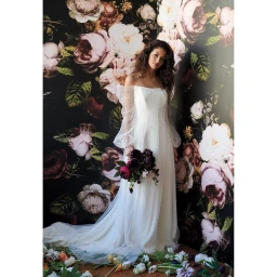 Woman in white off-the-shoulder gown holding a dark floral bouquet against a dark floral backdrop