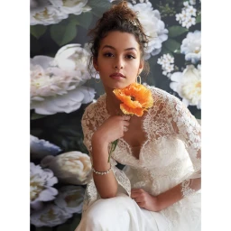 Woman in white lace top holding an orange poppy flower in front of a floral wallpaper backdrop