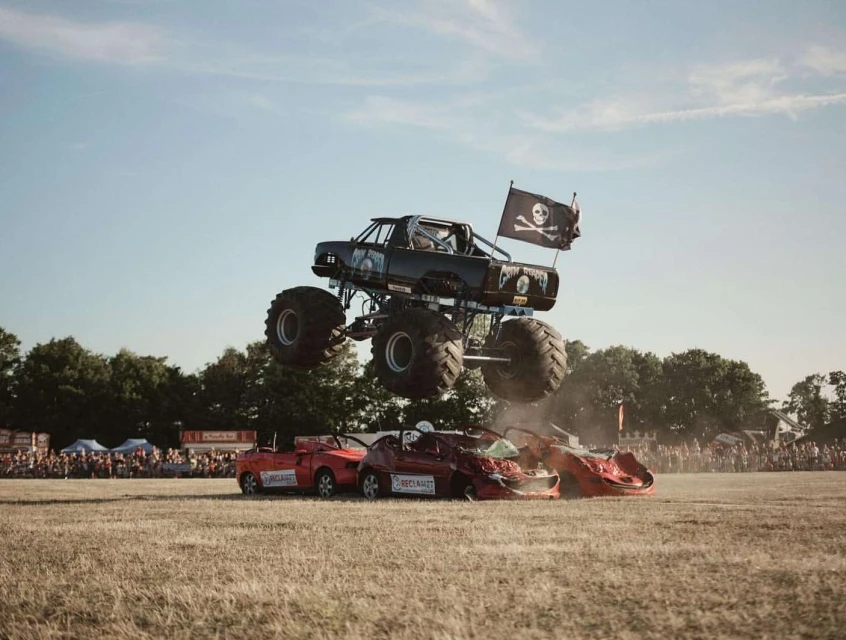Monster truck jumps over crushed cars during an outdoor daytime rally event with a crowd of spectators and a bright, clear sky.