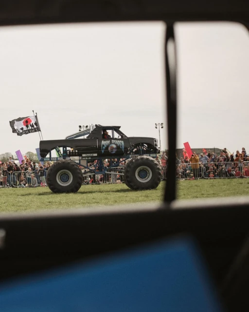 Monster truck event: A black monster truck on a grassy field is viewed through a vehicle window, with a crowd in the background.