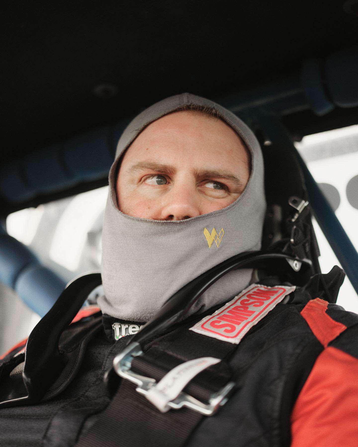 Man wearing a grey balaclava and a black and red racing suit, looking left while sitting inside of a vehicle cabin.