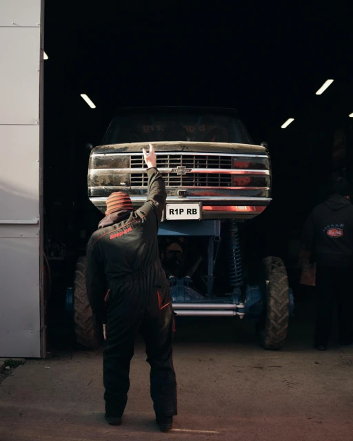 Mechanic in work clothes gesturing toward a lifted black monster truck in a garage with open door and concrete flooring.