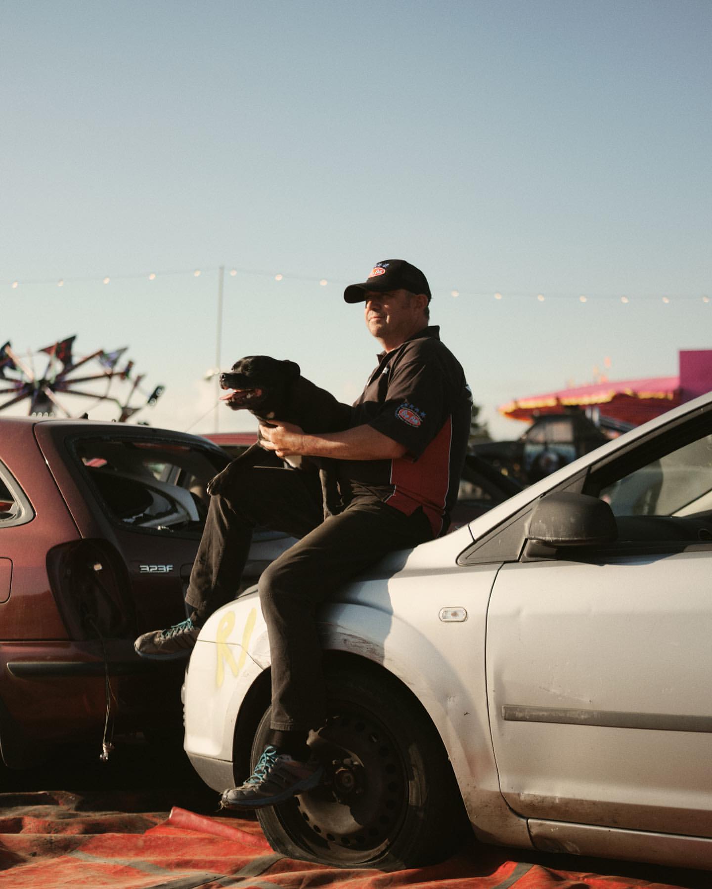 Man sits on damaged car with a dog, wearing cap and work shirt, posing in outdoor setting with blurred background.