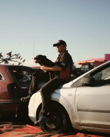 Man sits on damaged car with a dog, wearing cap and work shirt, posing in outdoor setting with blurred background.