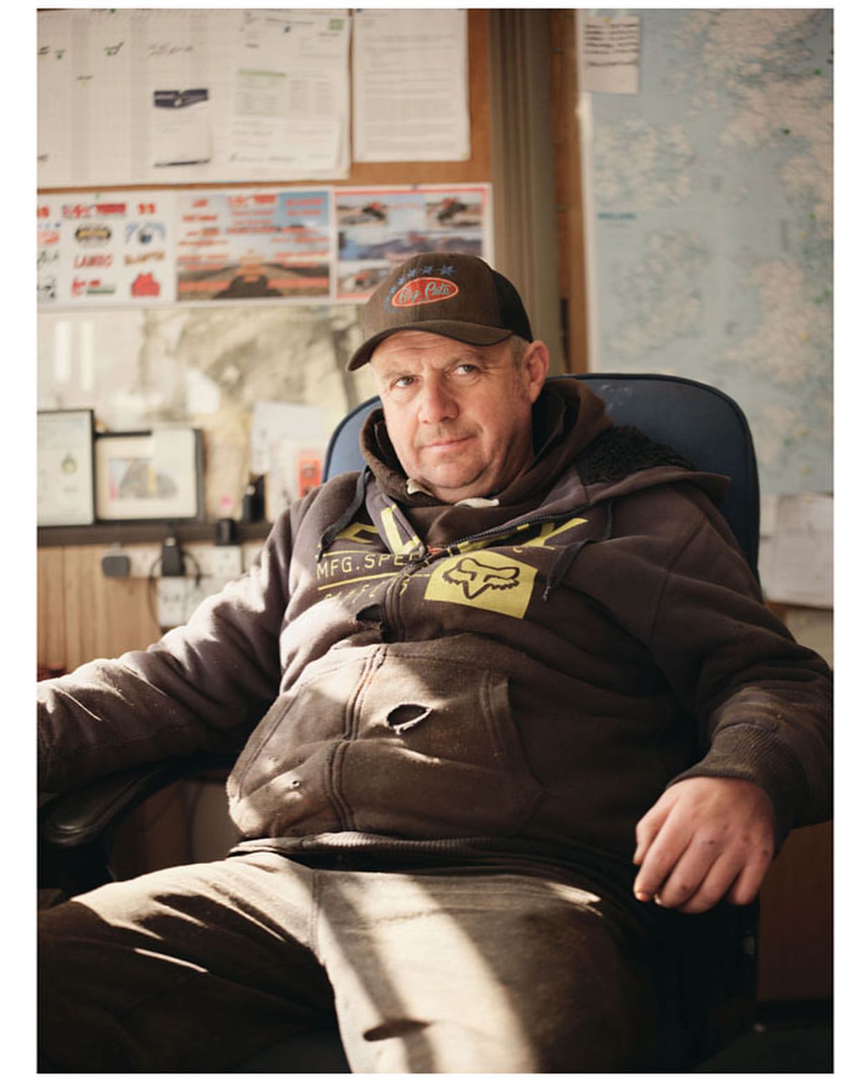 Man sits in a blue chair inside an office with papers and a map in the background, wearing a brown cap and a hooded sweatshirt.
