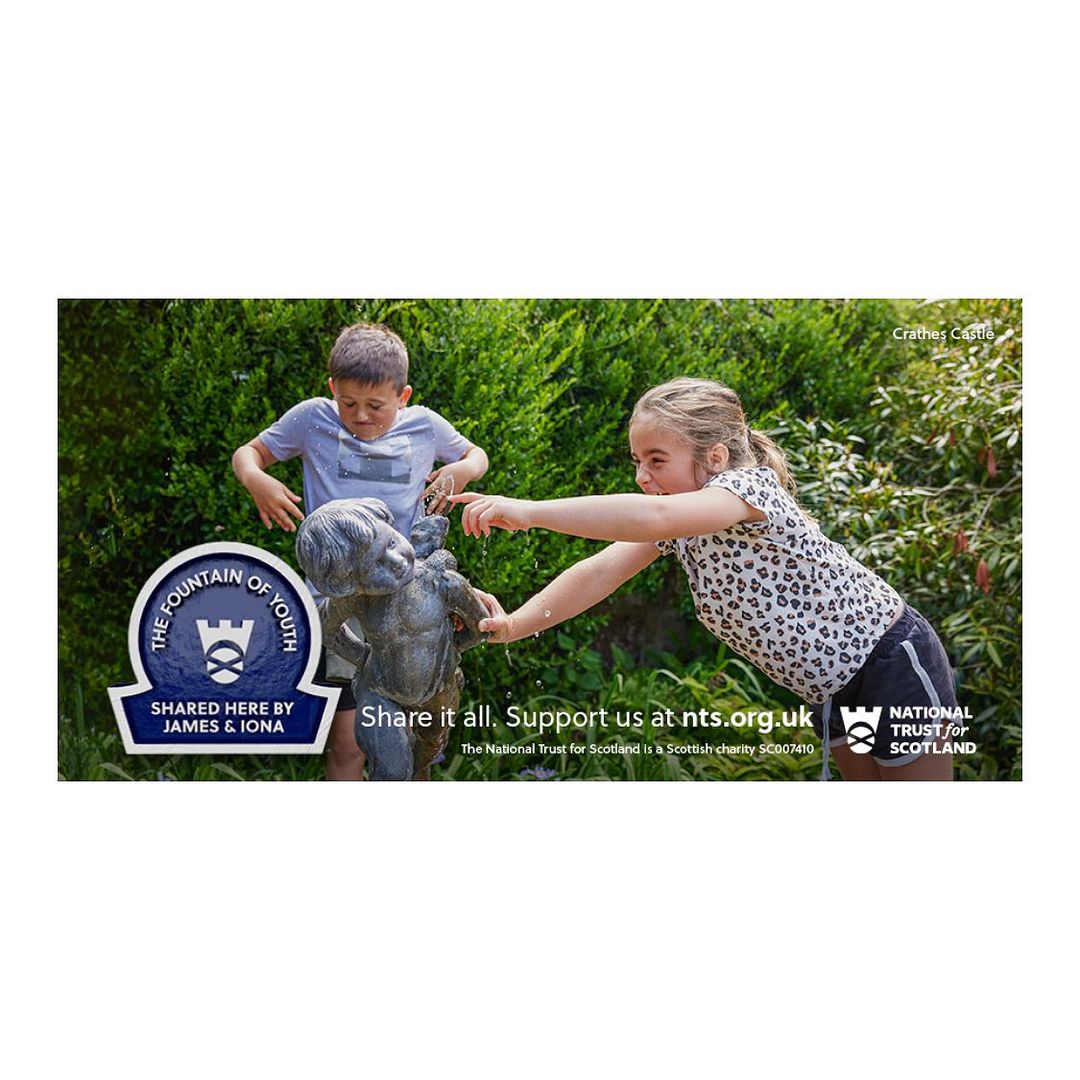 Two children, a boy and a girl, are playing with water from a statue in a garden setting, with green foliage in the background.