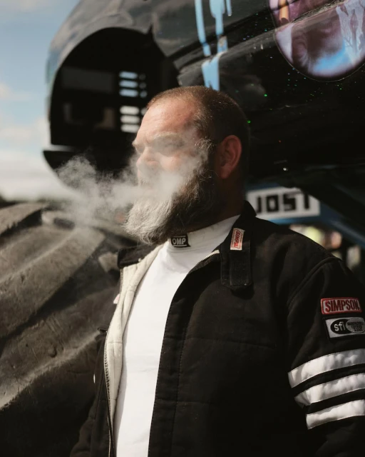 Man with a beard is smoking in front of a monster truck in an outdoor setting, wearing a racing jacket and a white undershirt.