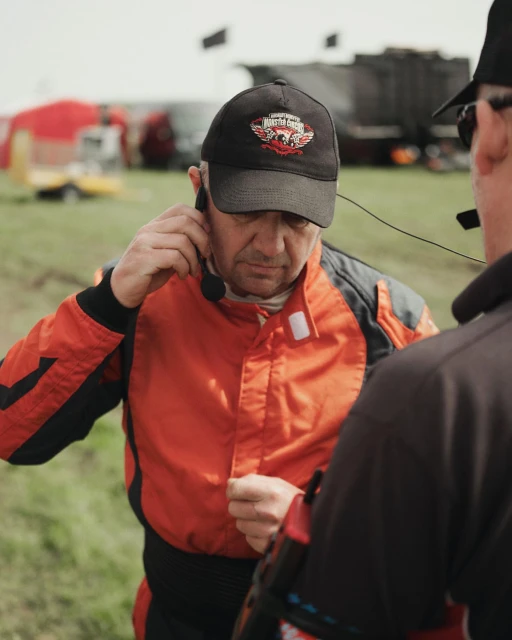 Two men wearing racing suits and headsets are standing outdoors with a trailer and other vehicles visible in the background on grass.
