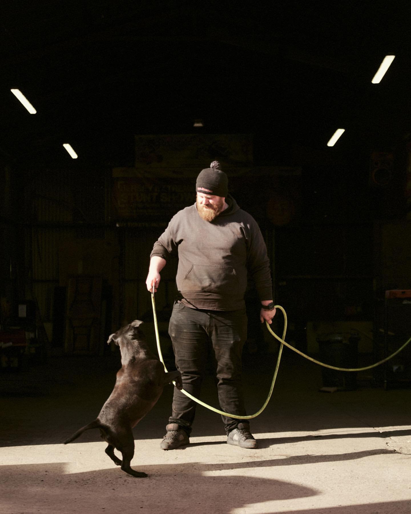Adult male trains black dog indoors, holding rope in industrial setting with concrete floor and natural, overhead fluorescent lighting.