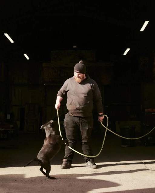 Adult male trains black dog indoors, holding rope in industrial setting with concrete floor and natural, overhead fluorescent lighting.