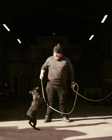 Adult male trains black dog indoors, holding rope in industrial setting with concrete floor and natural, overhead fluorescent lighting.