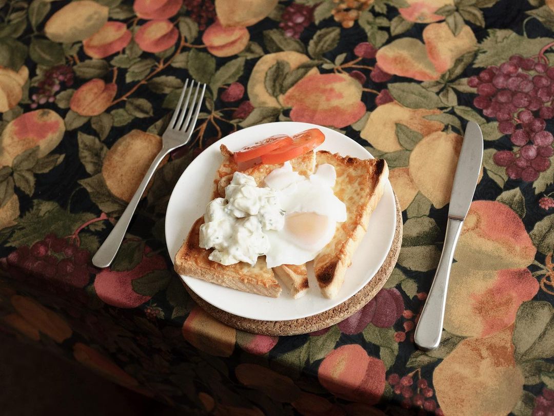 Breakfast meal with toast, poached egg, and tomato slices plated with fork and knife on floral tablecloth with dark background.
