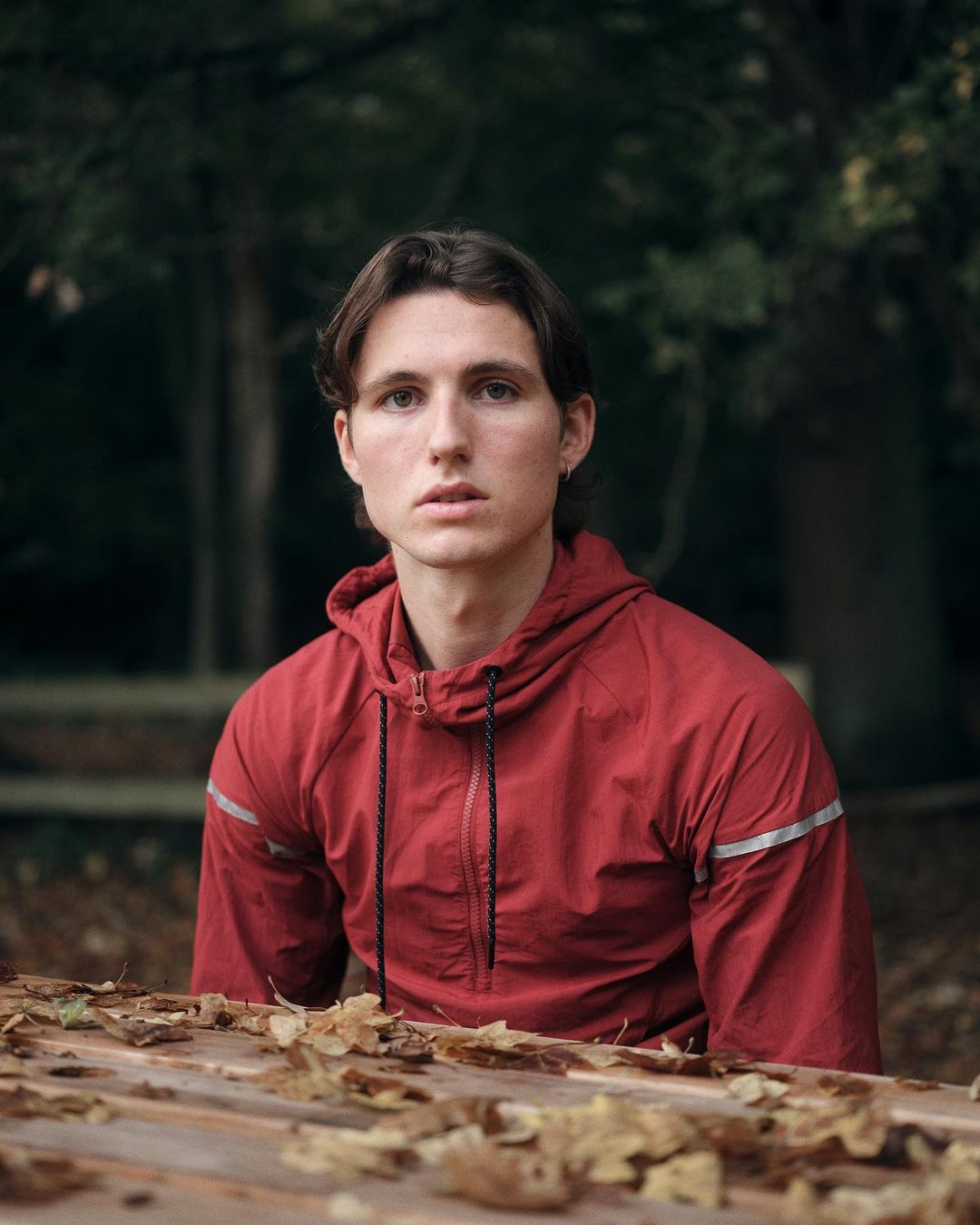 Portrait of a young adult man with medium-length brown hair in a red hooded jacket posing outdoors on a wooden surface covered with leaves.