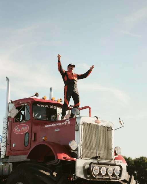 Adult male in a racing suit stands atop a red semi-truck, arms raised in victory against a clear sky backdrop, promoting Big Pete.