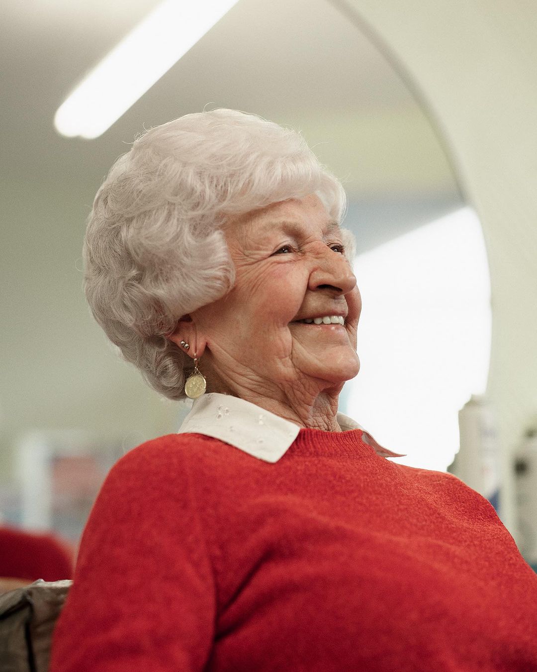 Close-up of a smiling elderly woman with short white hair wearing a red sweater in a bright indoor setting with a glimpse of a mirror.