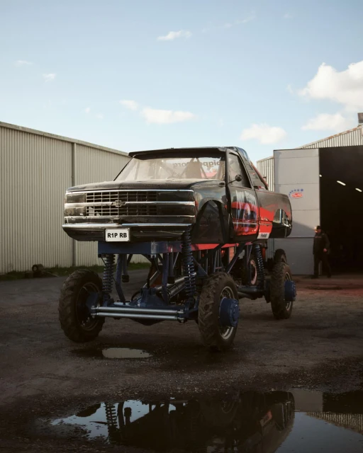 Modified monster truck on large tires stands in front of a garage with a person standing in the background during the daytime.