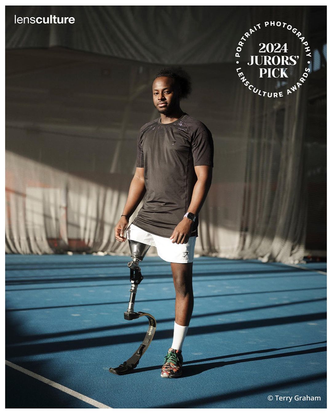 A full shot of a man with a prosthetic leg stands on an indoor court in athletic wear with soft natural lighting and a shallow depth of field.