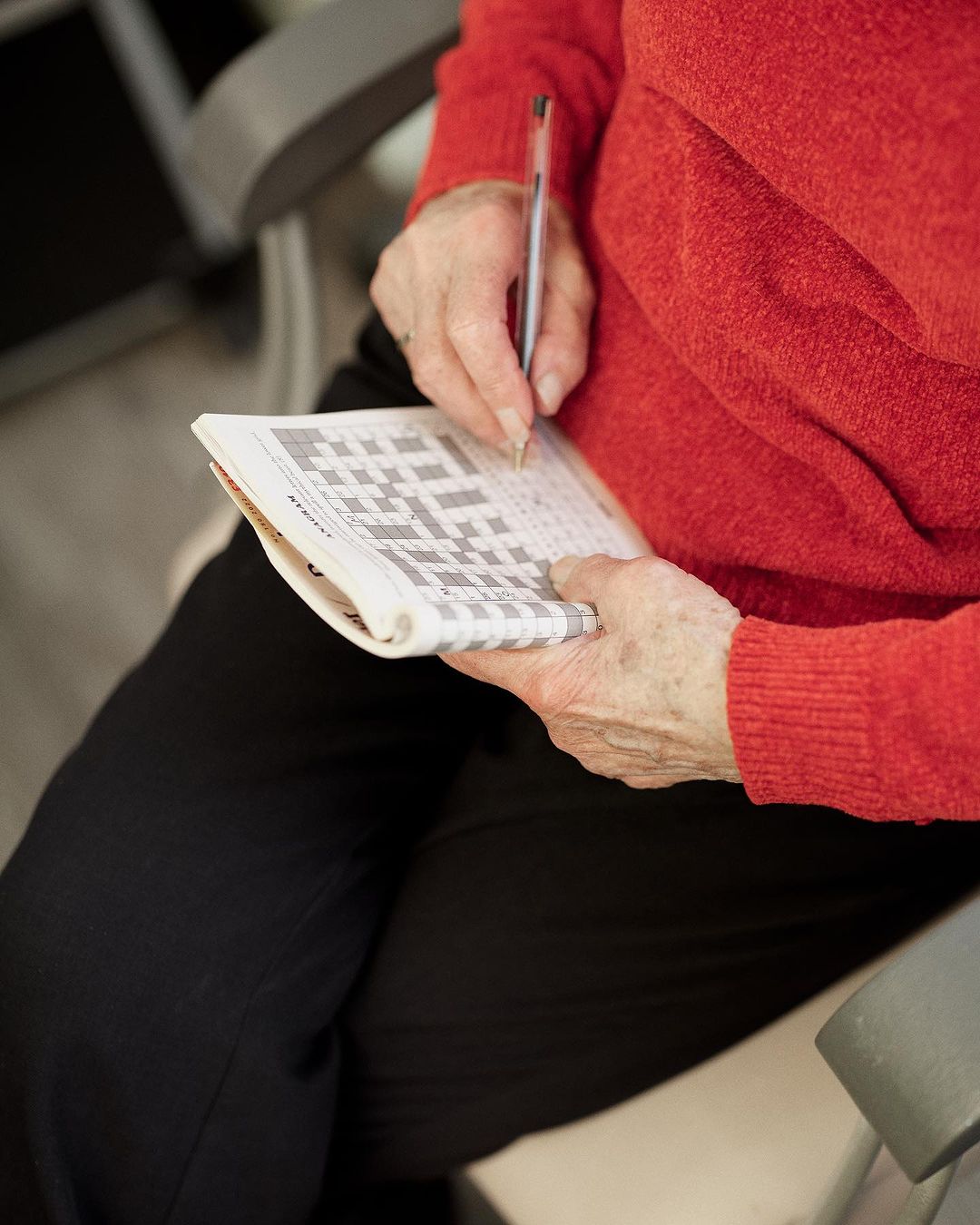 Elderly woman in red sweater seated, holding a pen and working on a crossword puzzle in an indoor setting with neutral colors.