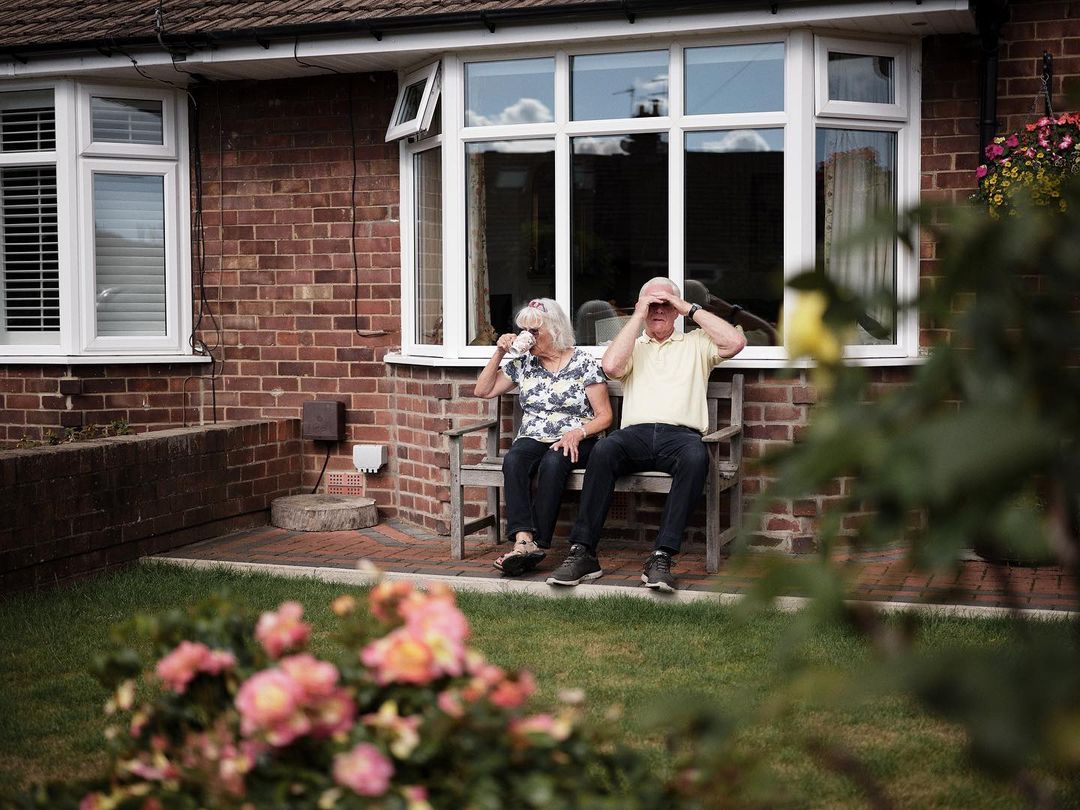Elderly couple relaxes outdoors, enjoying drinks on a wooden bench in front of a brick house, framed by lush garden foliage.