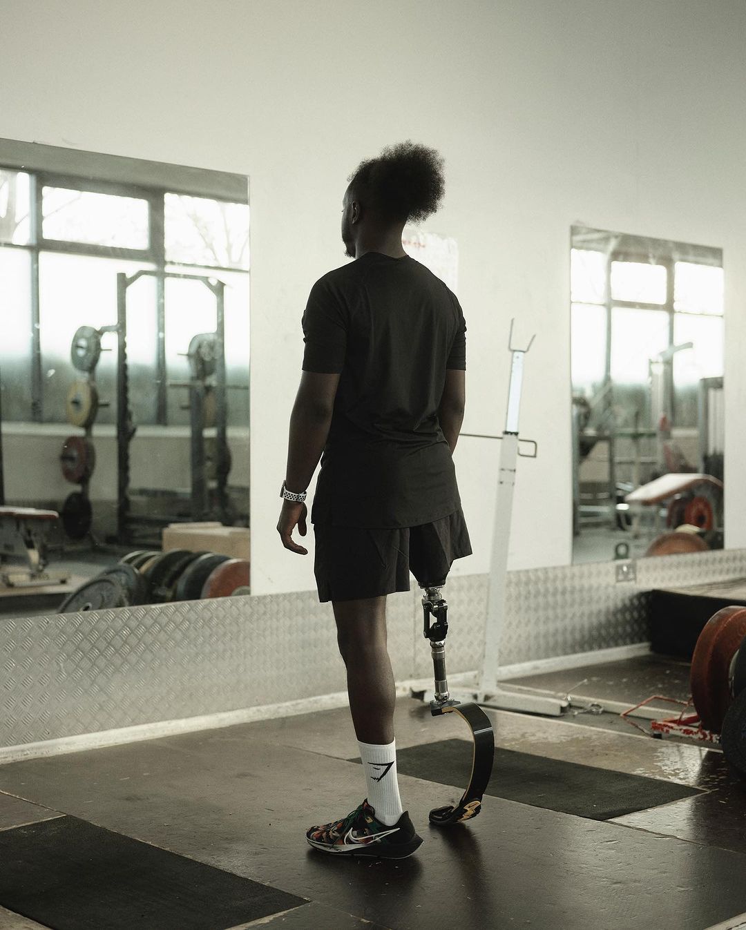 Man with prosthetic leg stands in a gym setting, facing a mirror with weightlifting equipment reflected in the background.