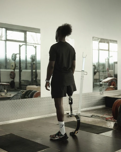 Man with prosthetic leg stands in a gym setting, facing a mirror with weightlifting equipment reflected in the background.