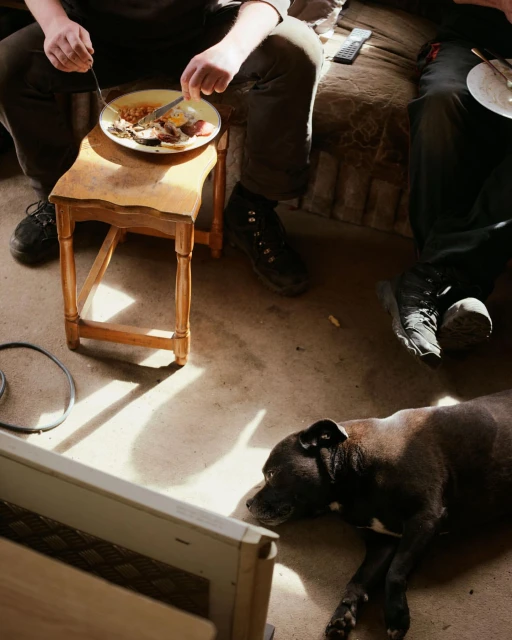 Two men eating while a dog lies in the foreground. A stool acts as a table with a plate of food and eating utensils visible.