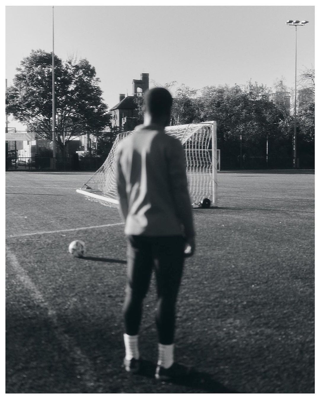 Monochrome image of an adult male standing on a soccer field with a soccer ball and goal net visible in the background.