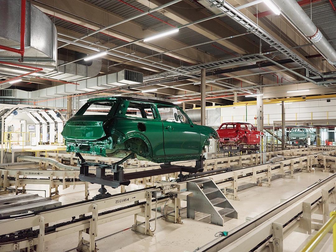 Automotive factory assembly line with green car bodies in progress, showing machinery, lighting, and stages of vehicle construction in a large space.