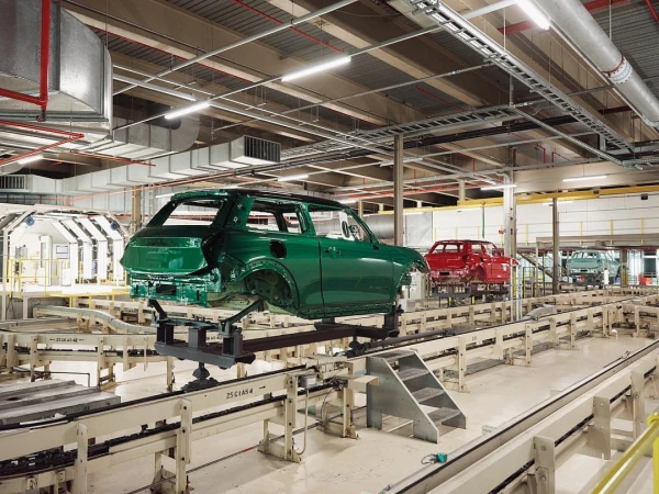 Automotive factory assembly line with green car bodies in progress, showing machinery, lighting, and stages of vehicle construction in a large space.