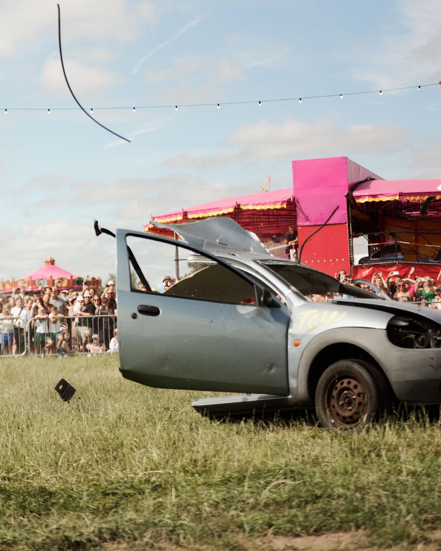 Damaged car with open door sits on grass with crowd and pink structure in the background under sunny sky, auto wreckage.